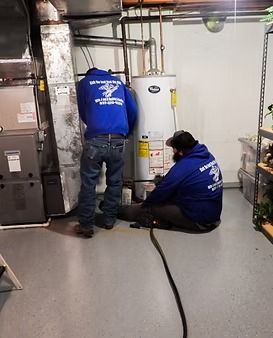 Two technicians in blue uniforms working on a water heater in a basement utility room