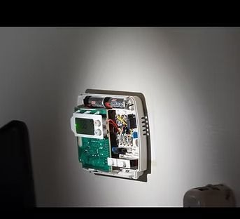 Open wall-mounted electronic device with exposed circuit board and wiring on a gray wall.