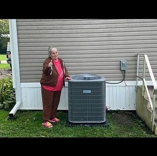 Woman standing beside outdoor air conditioner unit, gesturing with a thumbs-up in a backyard.
