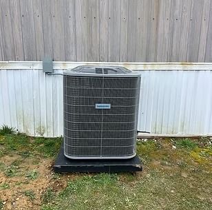 Outdoor air conditioner unit on a black pad against a white fence and wooden wall.