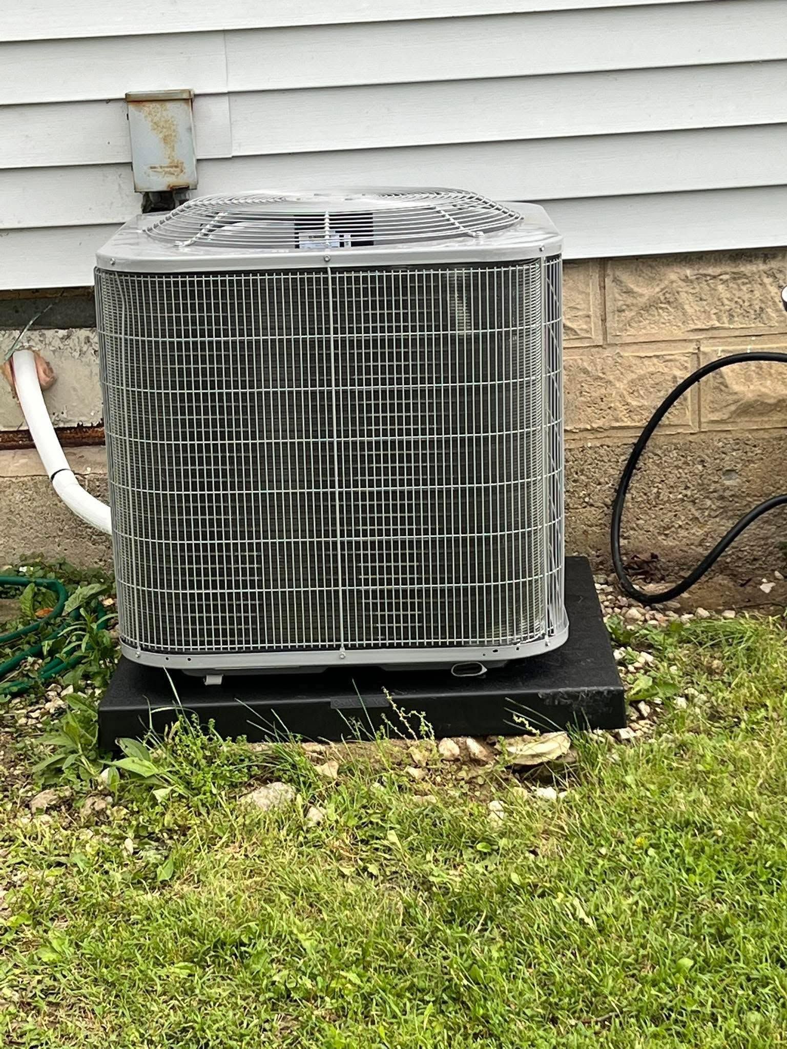 Outdoor central air conditioning unit beside a house with white siding and stone foundation.