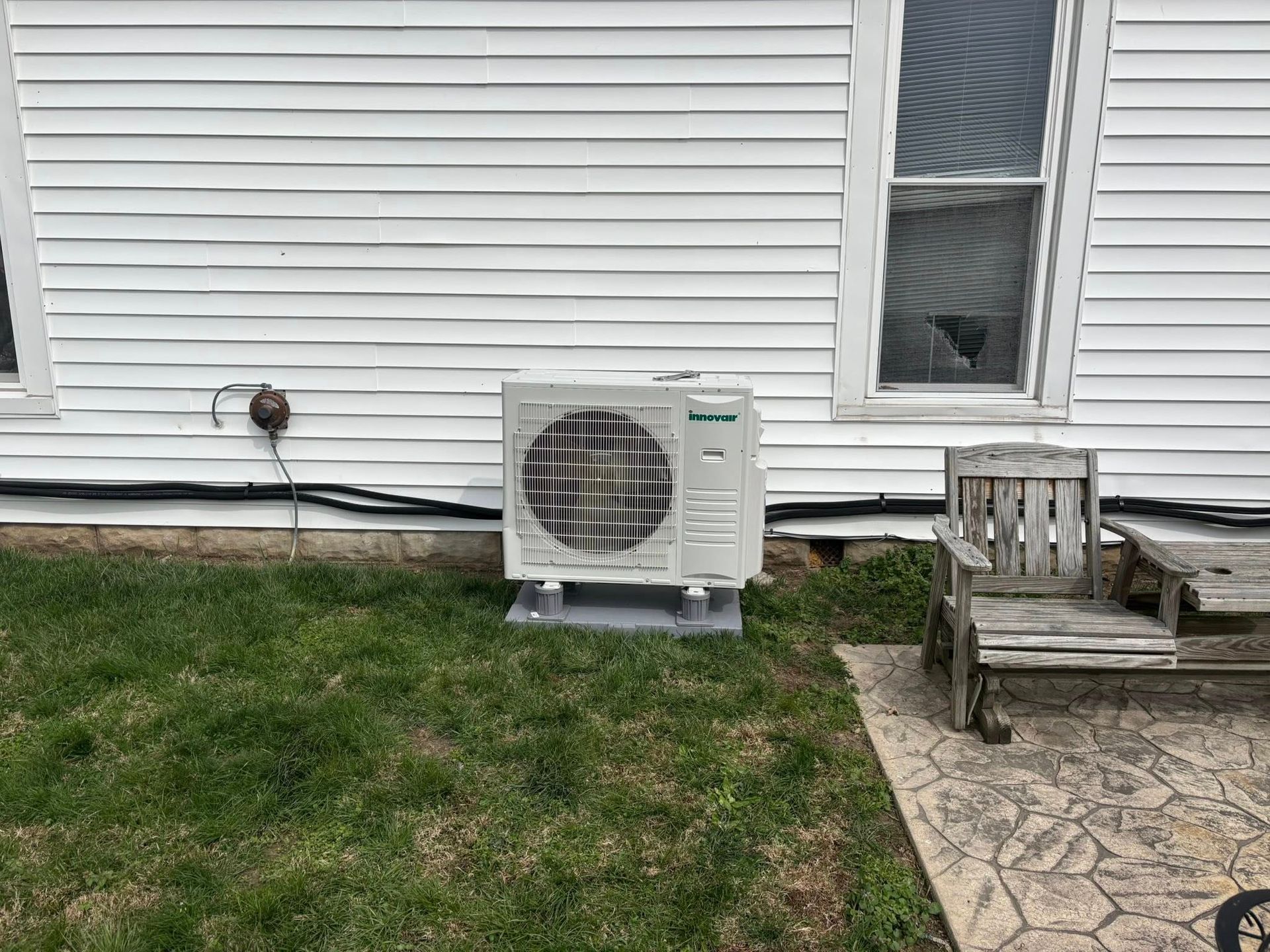Outdoor air conditioner unit against white house siding beside a wooden chair and stone patio.