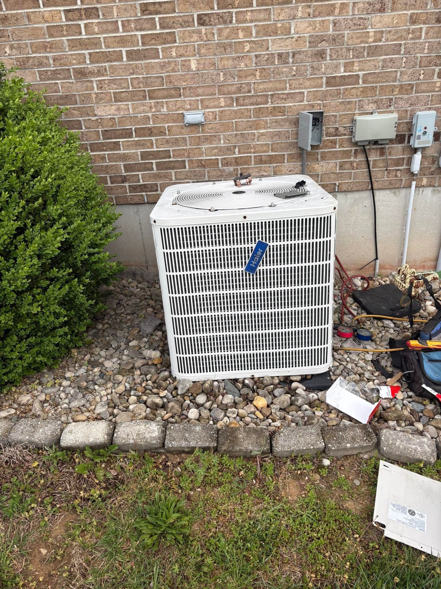 Outdoor HVAC unit beside a brick house, with shrubs, rocks, and electrical boxes nearby.