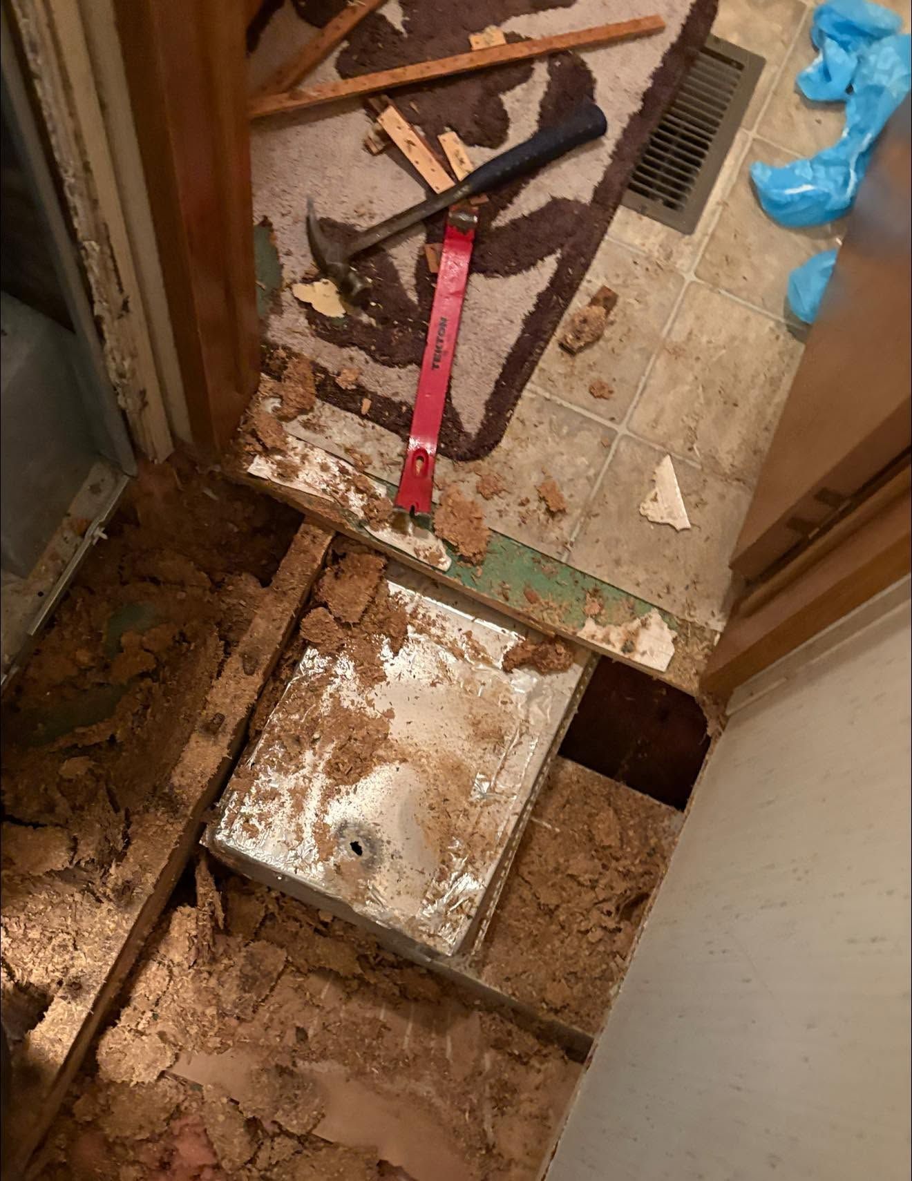 Messy bathroom floor with exposed subfloor, tools, dirt, and blue gloves during repair work