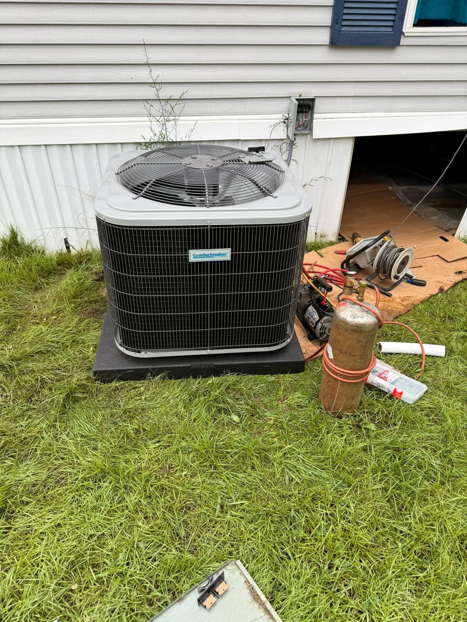 Outdoor air conditioner unit beside a house with tools and a rust-colored bottle on the grass