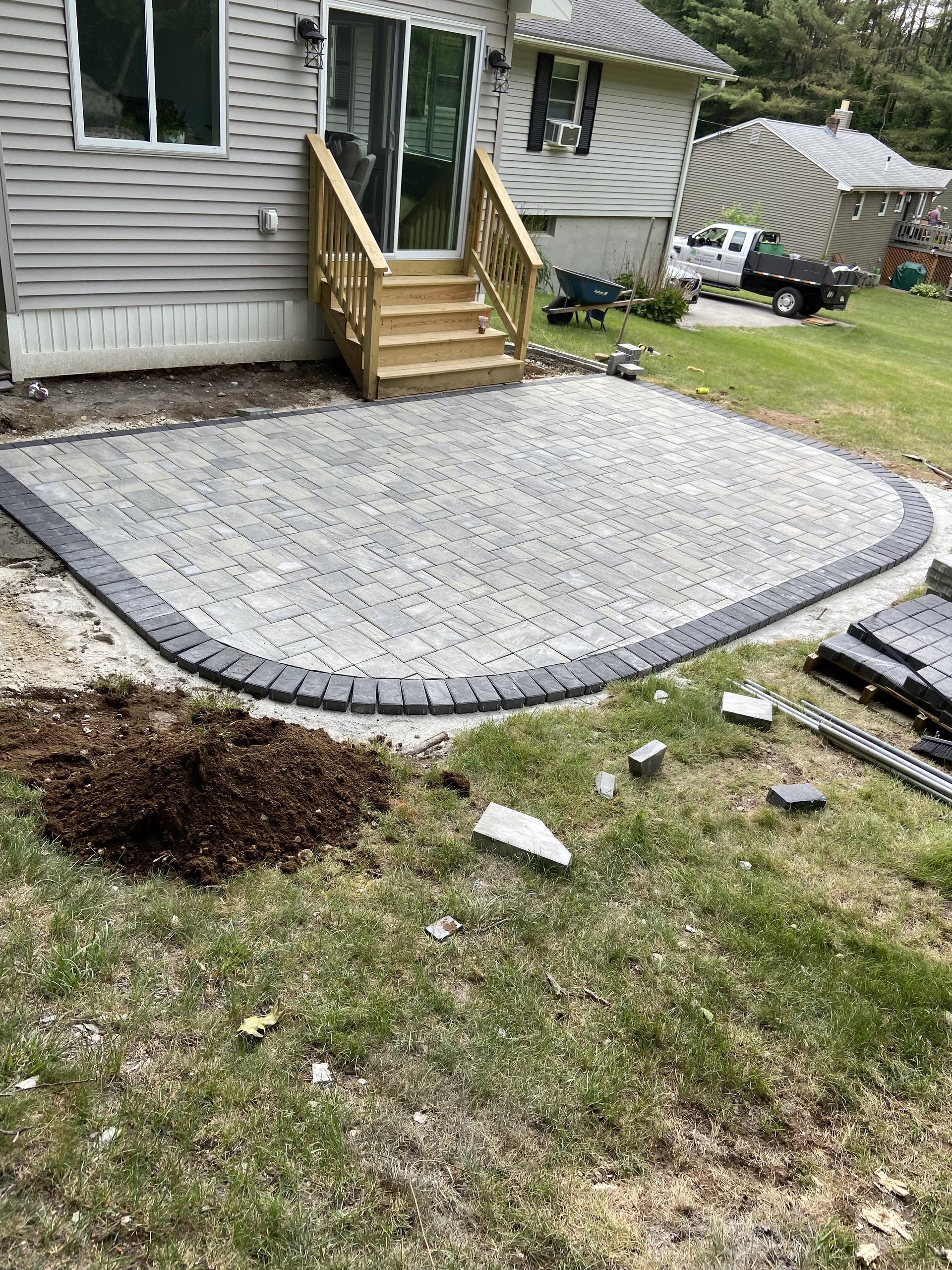 Patio with gray pavers and dark border, next to a grassy yard and a house with wooden stairs.