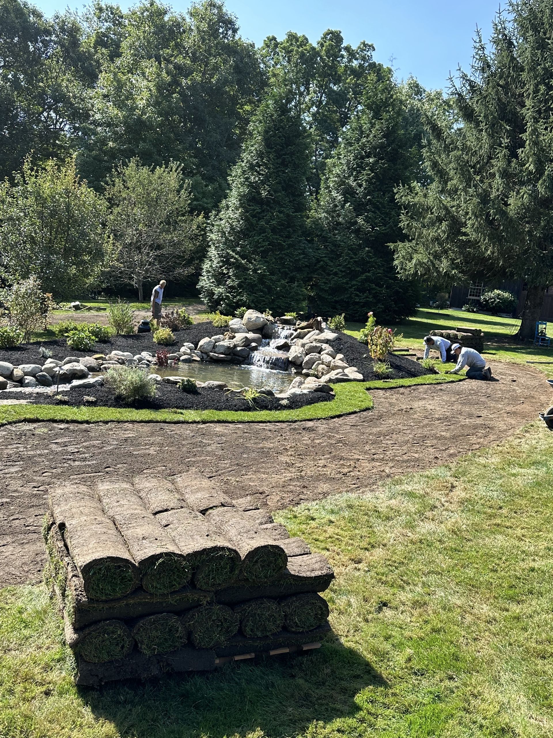 A stack of rolled sod in a park setting. Workers near a water feature and trees.