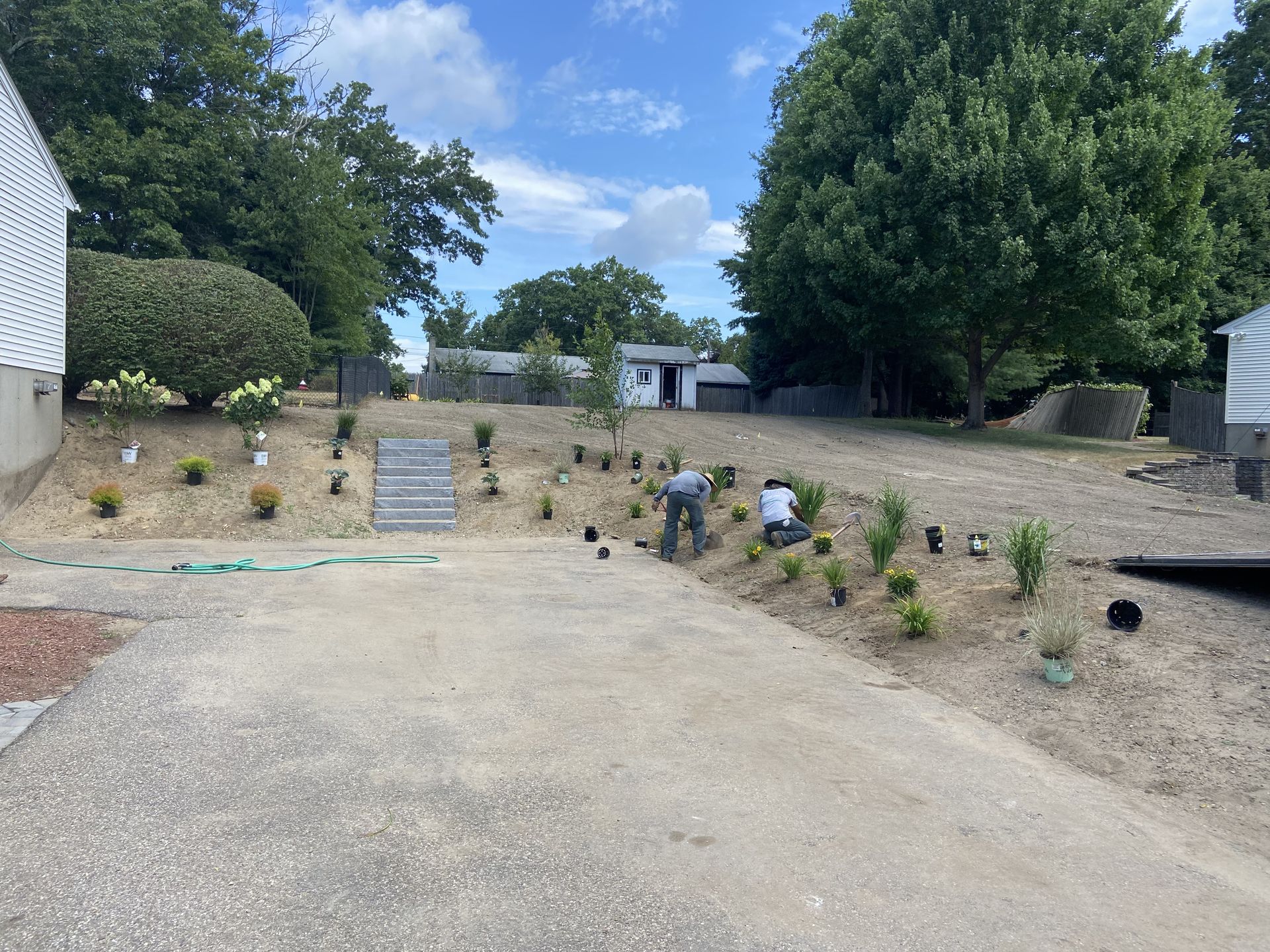 Two people planting on a sloped, gravelly area; a small white shed is visible in the background.