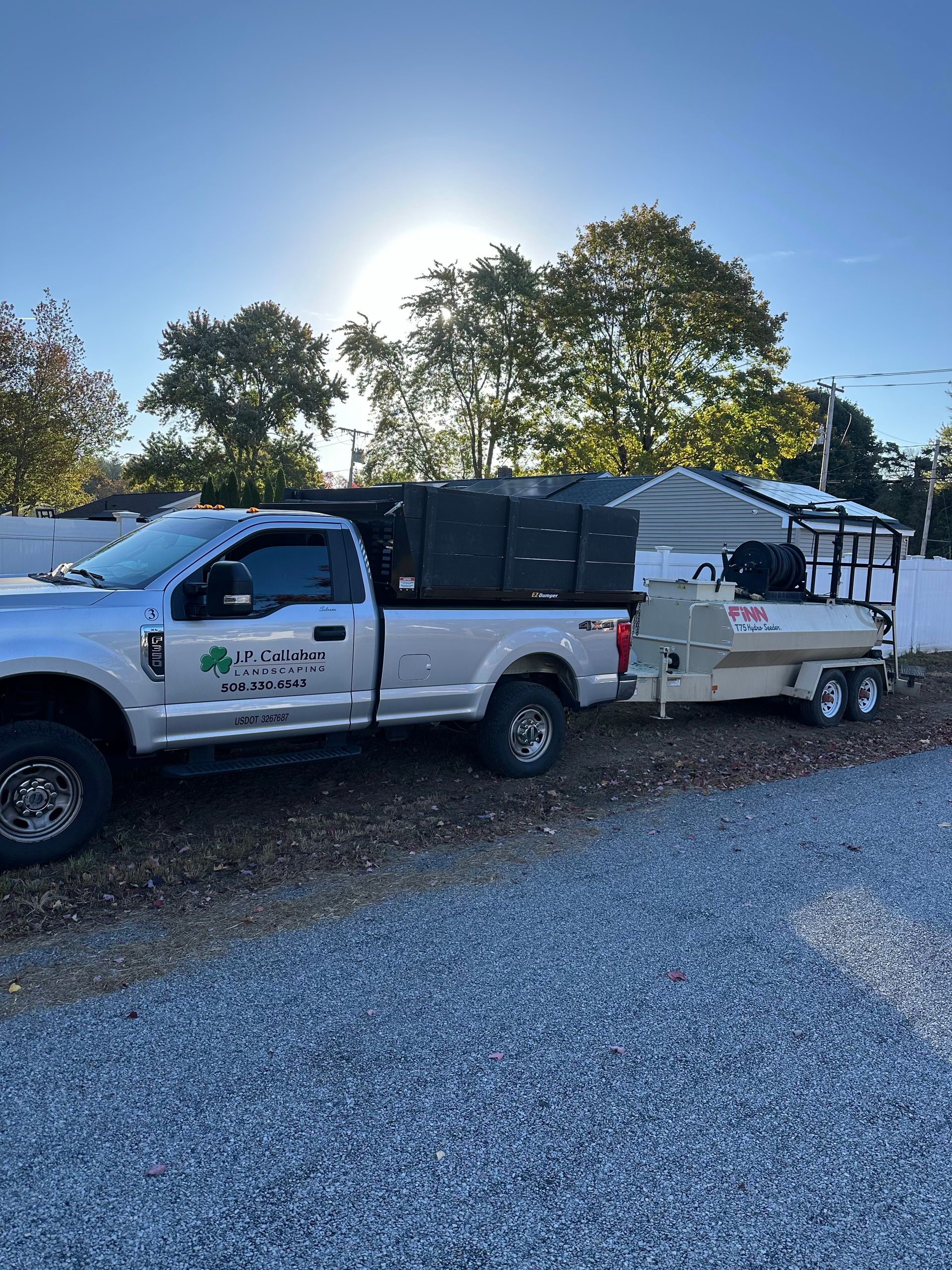 Silver pickup truck towing a trailer; both are parked on gravel. Sun is overhead, shining.