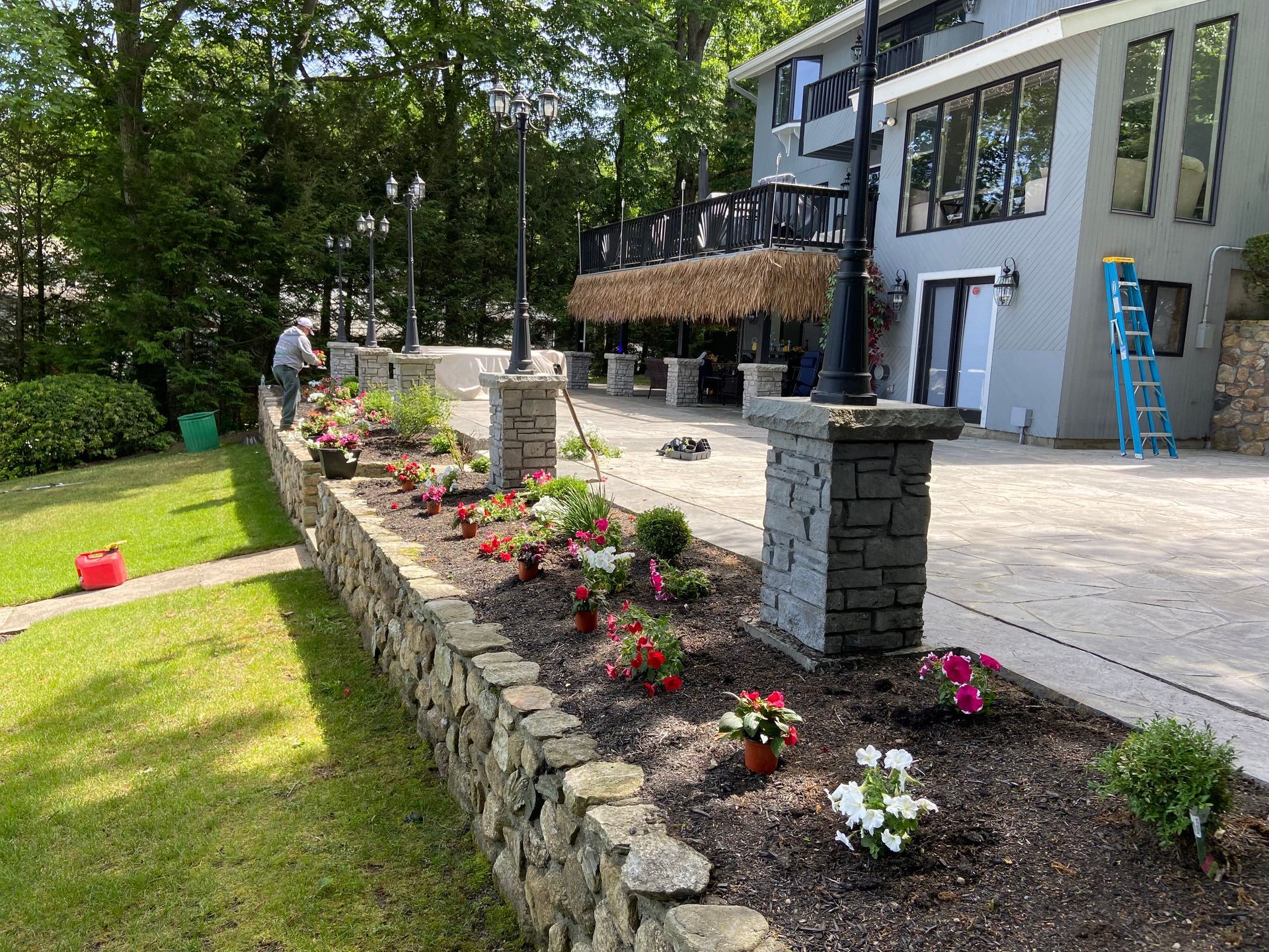 Stone wall with flower bed, lamp posts, house, and person gardening.