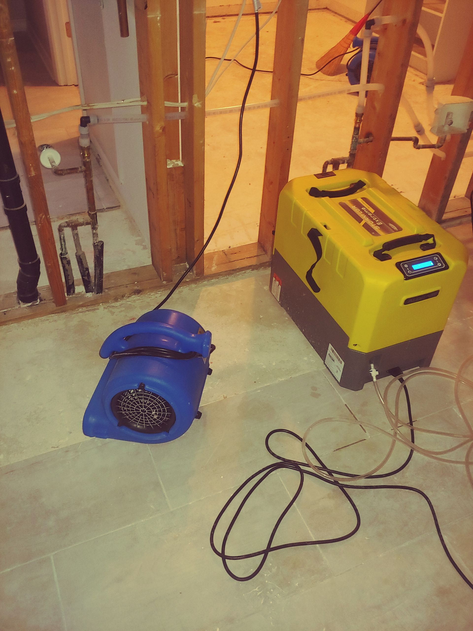 Blue air mover and yellow dehumidifier in a room with exposed framing, possibly a flood damage restoration.