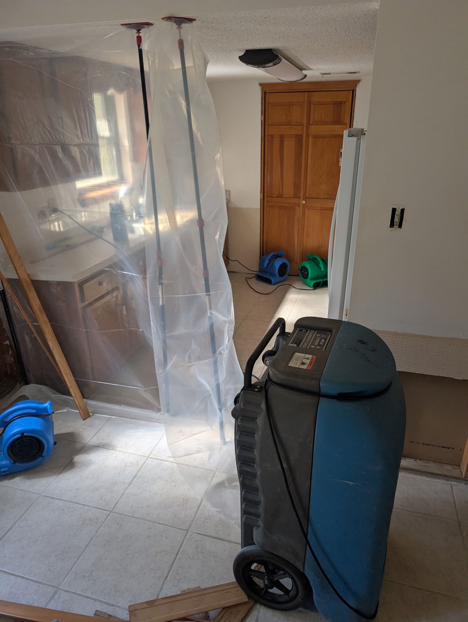 Dehumidifier and fans inside a room, plastic sheeting forming a barrier. Cabinets, window, and doorway visible.