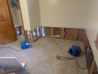 Room with damaged wall, exposed studs, and two blue fans blowing air on the concrete floor.
