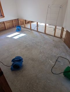 Room undergoing water damage restoration with exposed wall framing, concrete floor, and three fans.