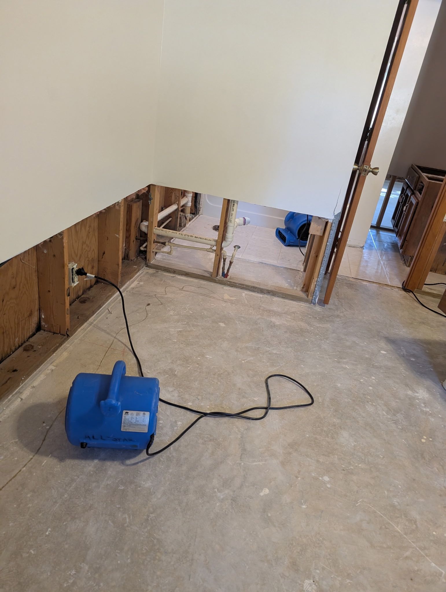 Blue fan on a concrete floor drying water damage. Exposed wall studs and plumbing visible.