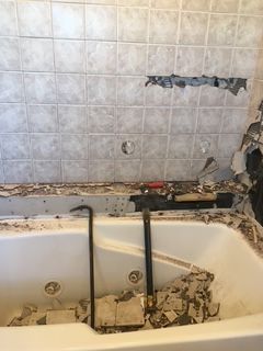 Bathroom tub with damaged tile wall and demolition debris. Crowbar and pipes are visible.