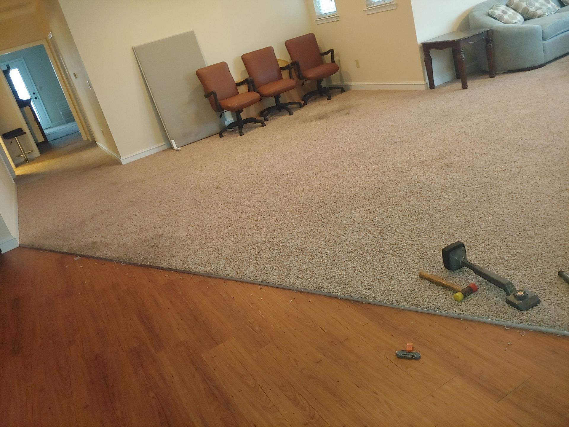 Beige carpet installed over wooden floor with three chairs and a sofa in the background.