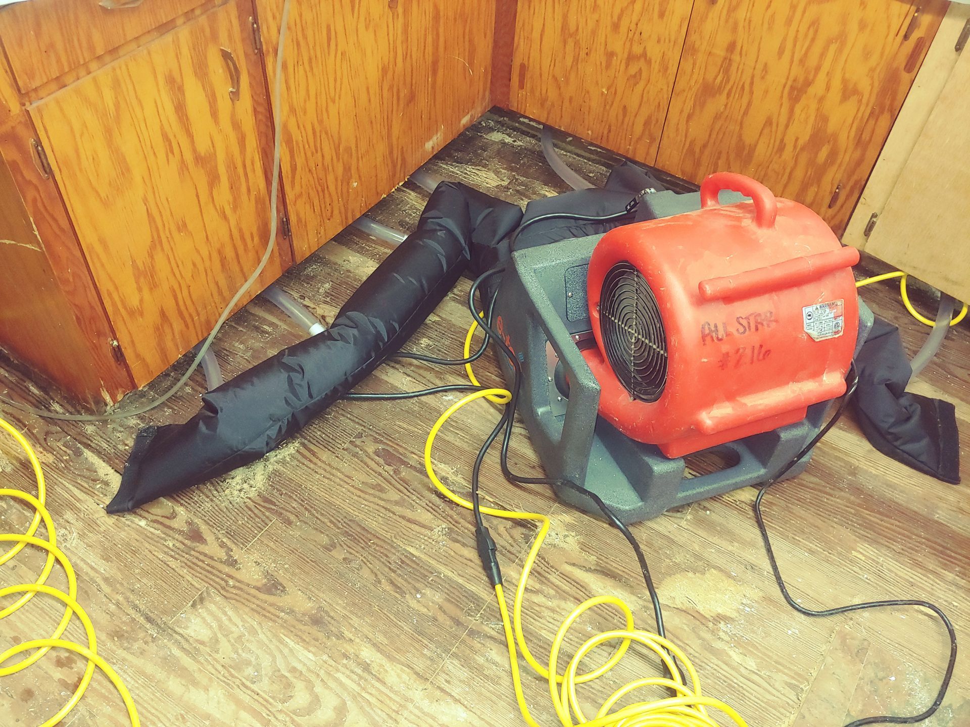 Red air mover with black duct in corner of room, yellow cords on floor.