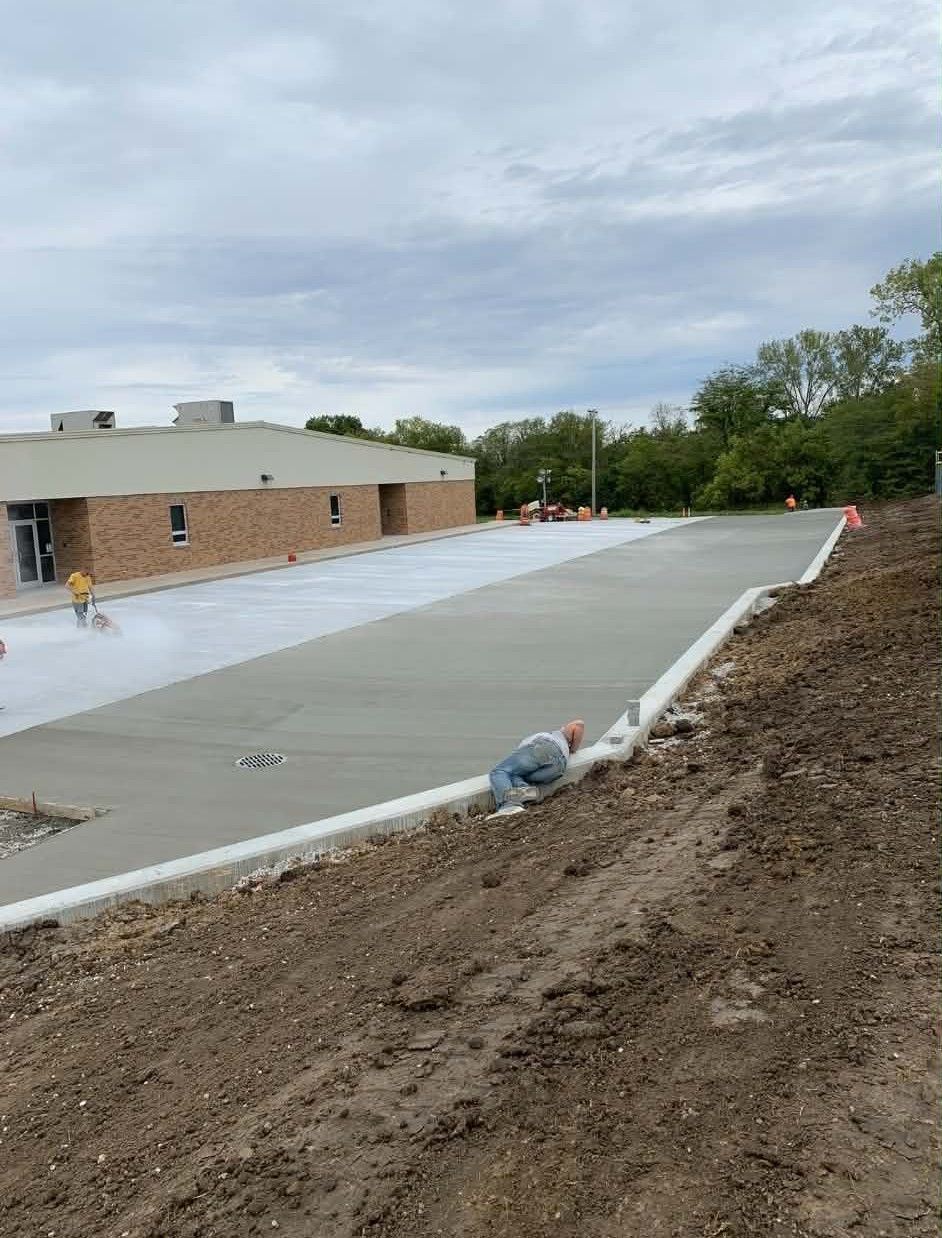 New concrete parking lot beside a brick building, with dirt grading and orange cones along the edge