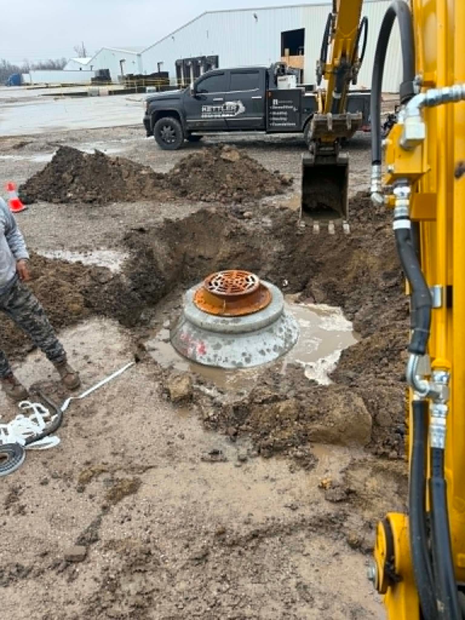 Construction site with excavator digging around a buried concrete manhole in muddy ground.