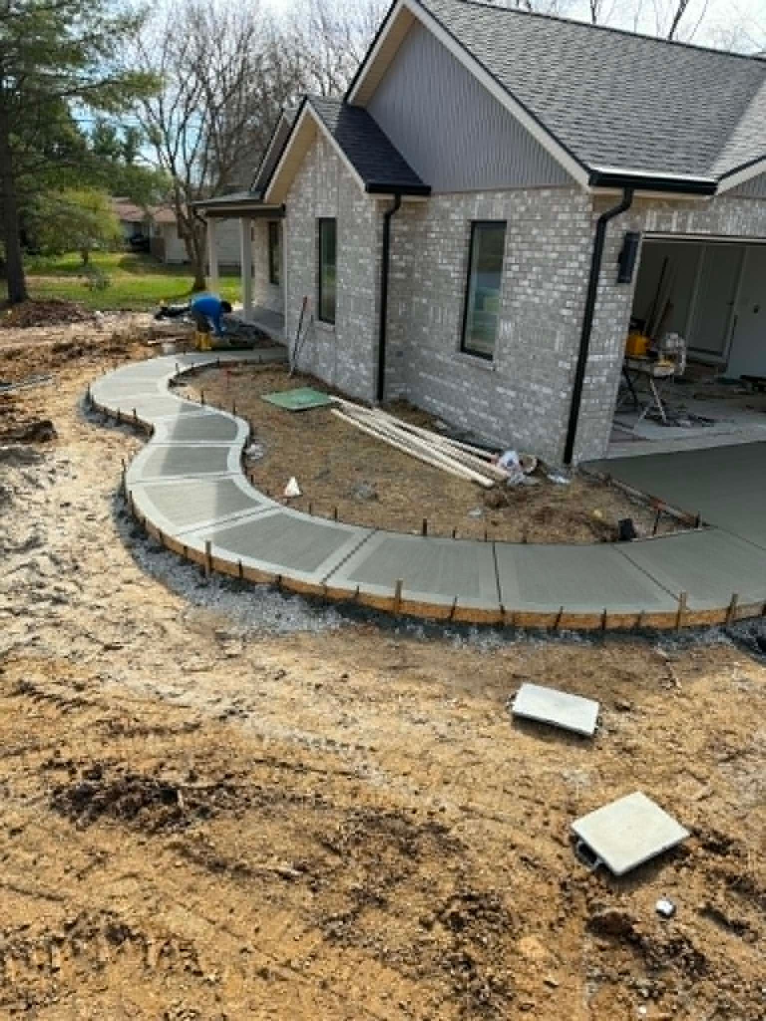 Front yard under construction beside a gray brick house, with a curved stone walkway being installed.