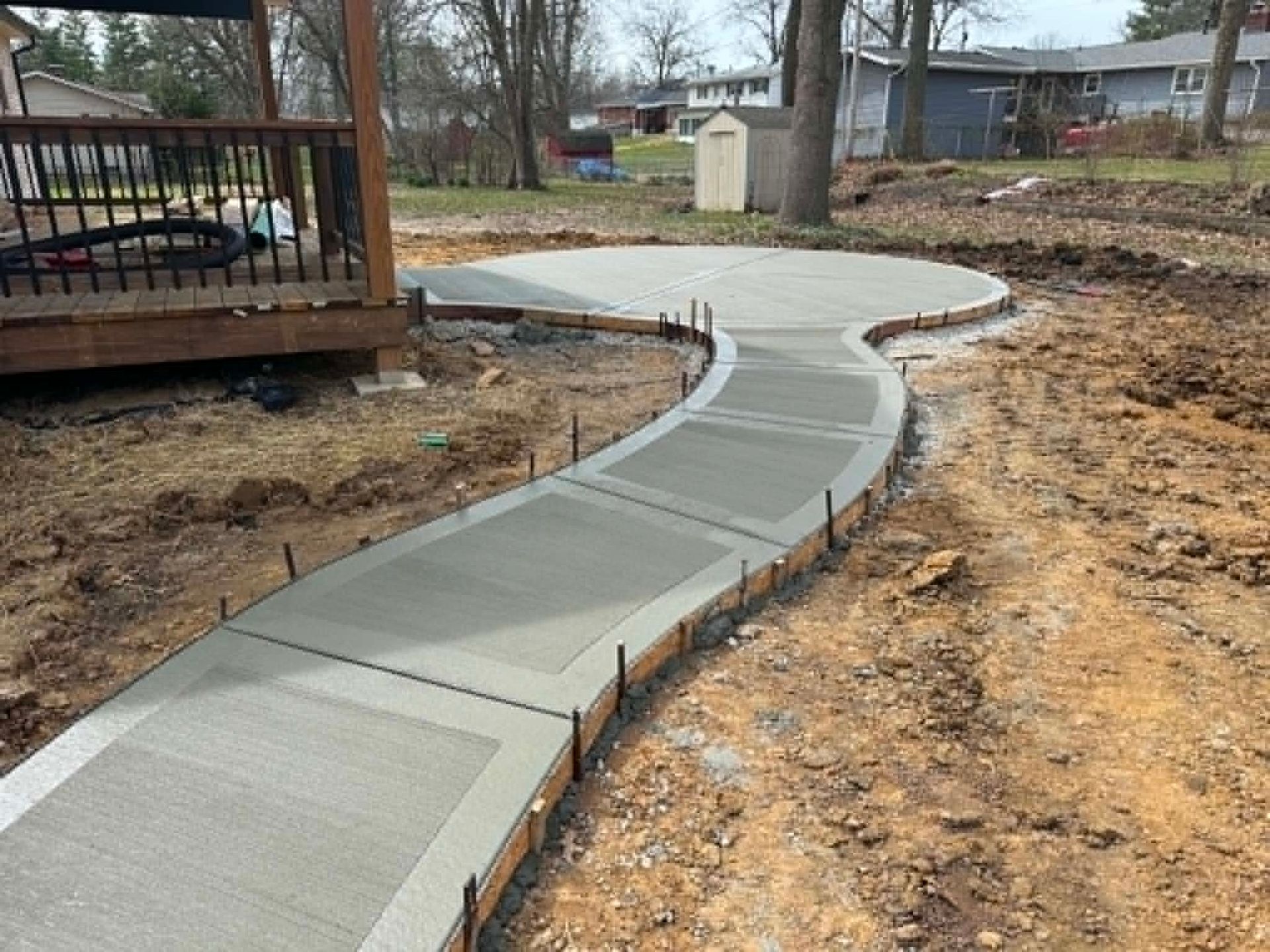 Curving concrete walkway under construction beside a wooden deck in a dirt yard.