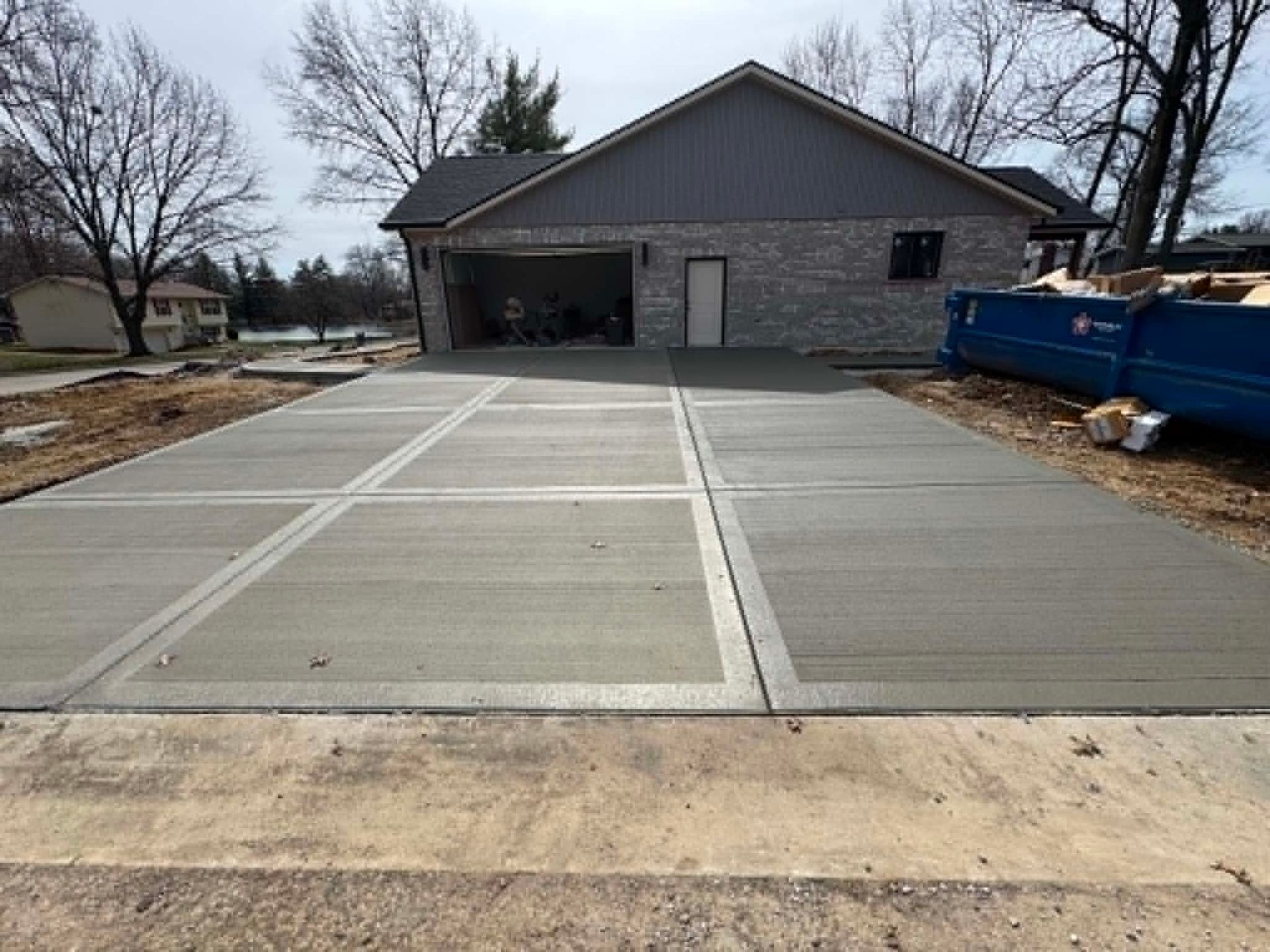 Concrete driveway with marked parking spaces leading to a gray building, with a blue dumpster on the right.
