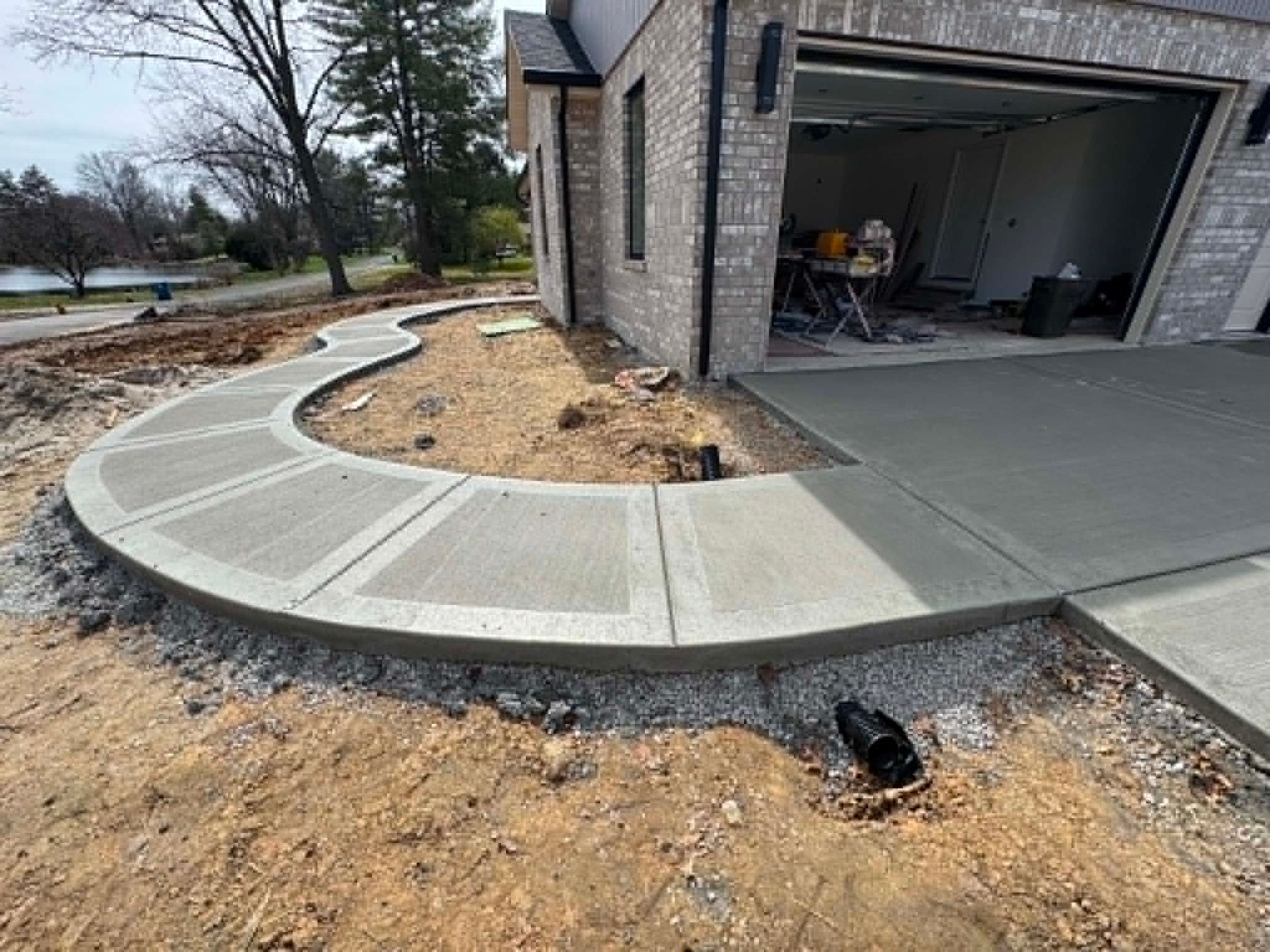 Curved concrete sidewalk and driveway beside a garage, with unfinished dirt landscaping and a drainage inlet.