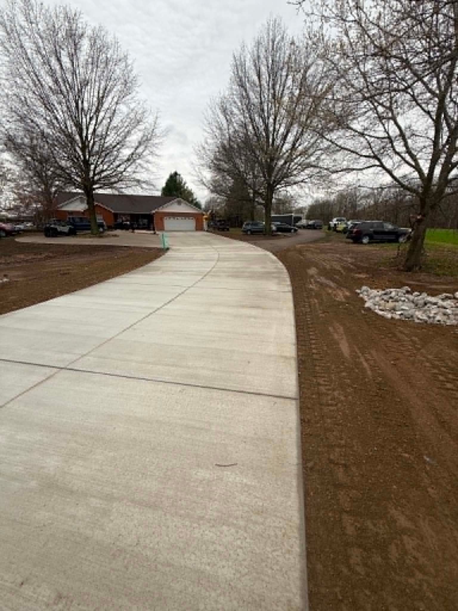Curved concrete sidewalk beside leafless trees, houses, and parked cars on a muddy roadside.