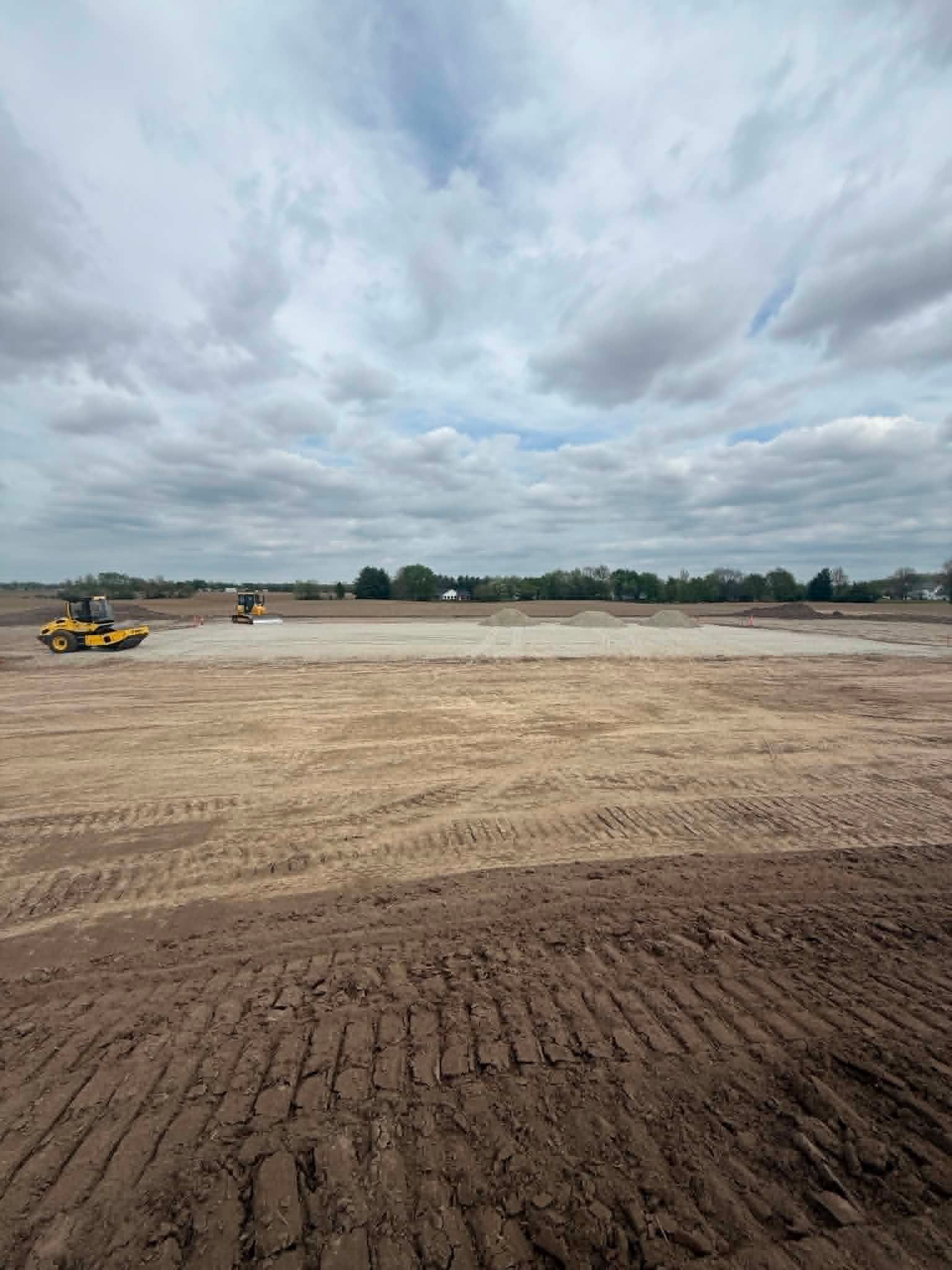 Wide dirt construction site under cloudy sky, with a yellow roller on the left and leveled soil in the foreground