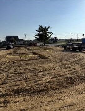 Empty dirt lot with tire tracks, a tree in the center, and cars parked along the road under a clear sky