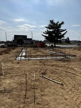 Construction site with foundation rebar in a sandy lot under a cloudy sky, trees and buildings in the background