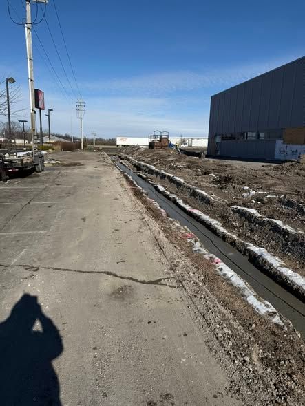 Cracked roadside beside muddy drainage ditch and a large blue building under a clear sky