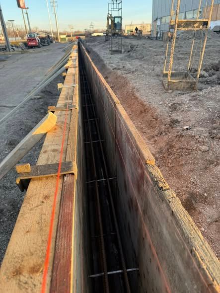 Long trench excavation beside a road, with rebar and wooden formwork at a construction site.