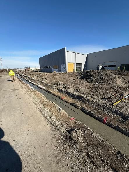 Construction site with trench along a dirt road beside a gray industrial building under blue sky