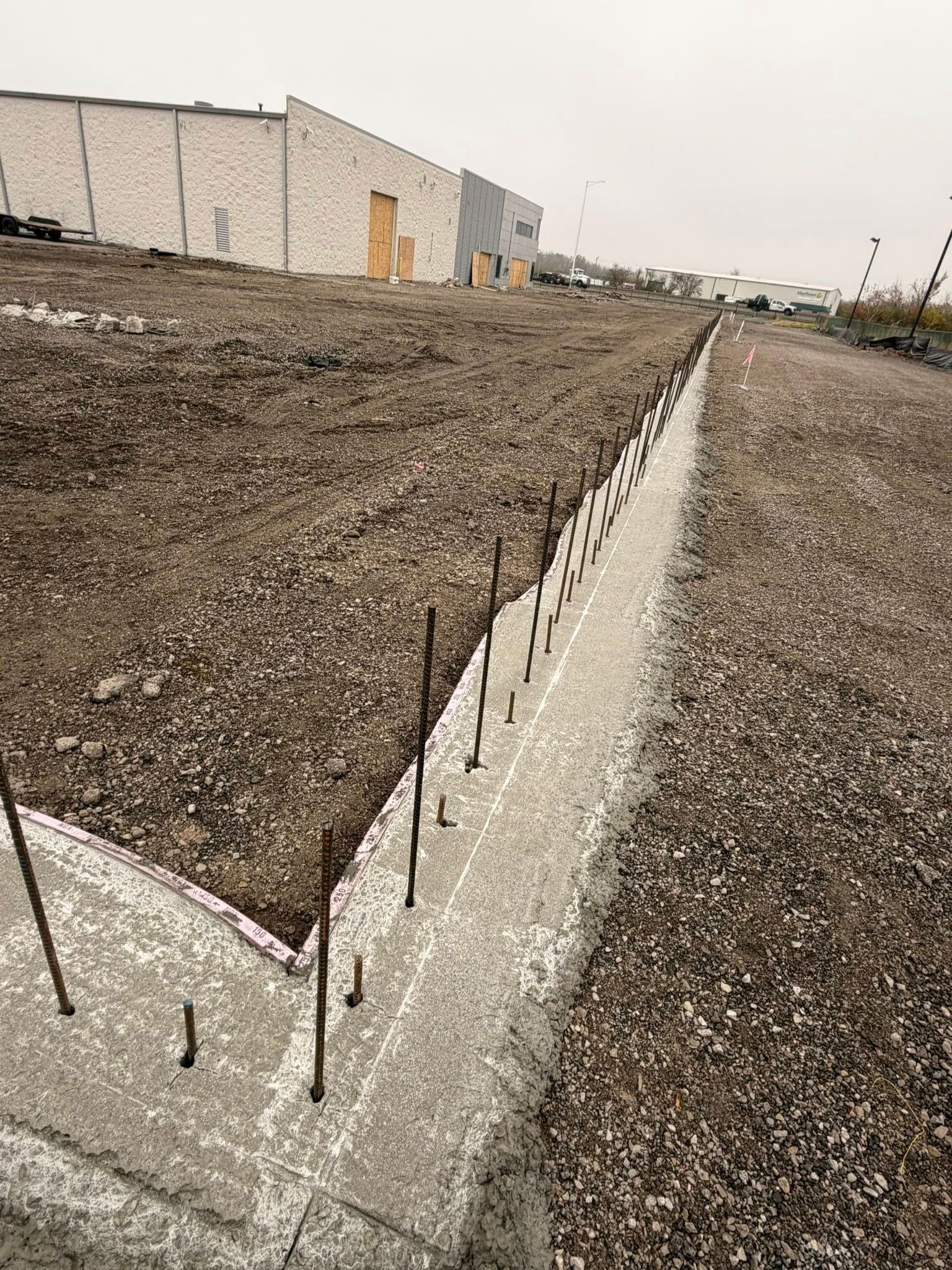Concrete footing with exposed rebar along a dirt lot beside a beige industrial building