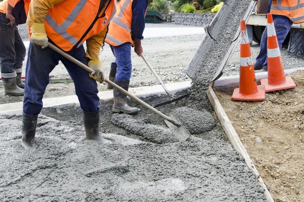 Workers in safety vests pouring and spreading wet concrete on a road construction site.