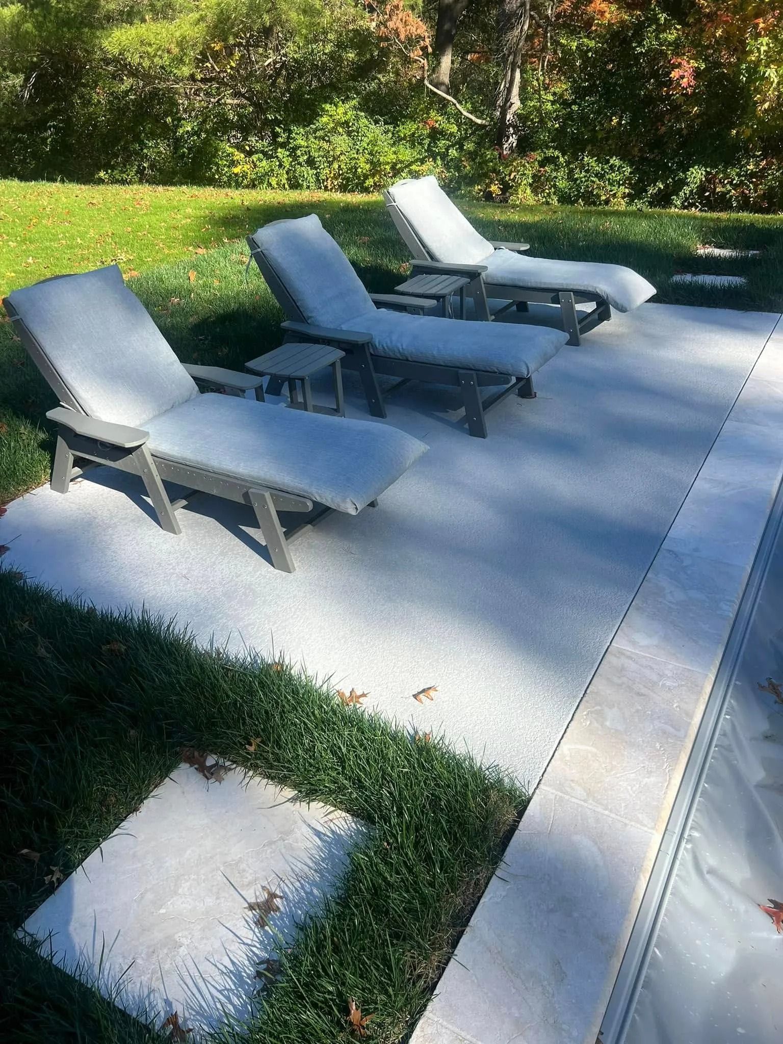 Three lounge chairs on a concrete patio beside a pool, set in a grassy yard.