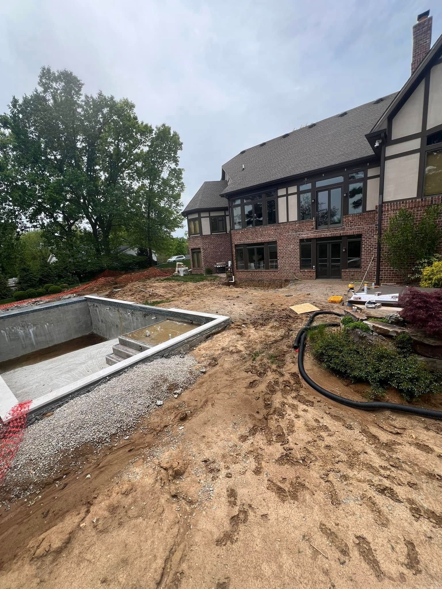Backyard with house and pool under construction; dirt, gravel, unfinished pool, brick house with large windows.