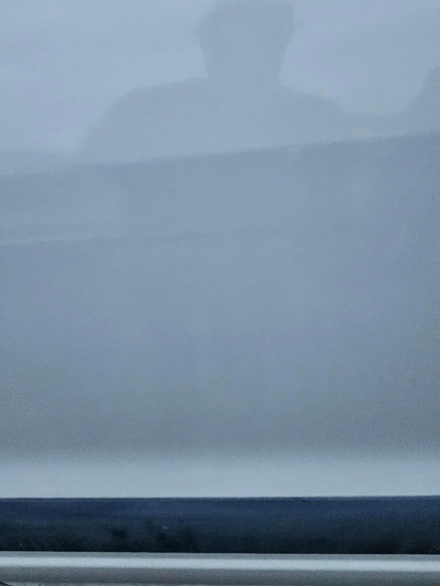 A reflection of a person in a window, with a dark, horizontal bar visible at the bottom against a blurred, gray background.