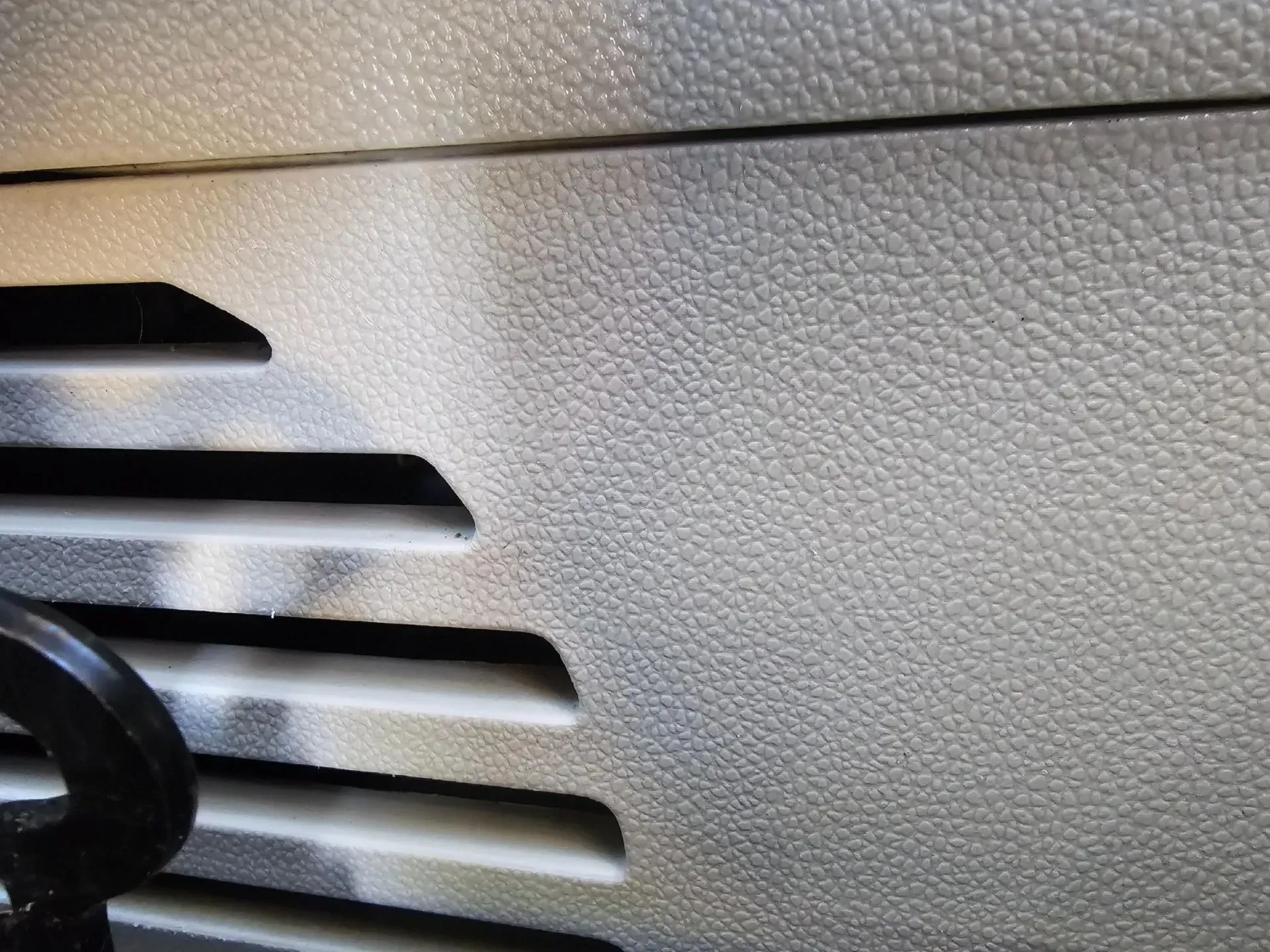 A close-up of a grey, textured plastic surface with horizontal ventilation slats and a black cylindrical component.