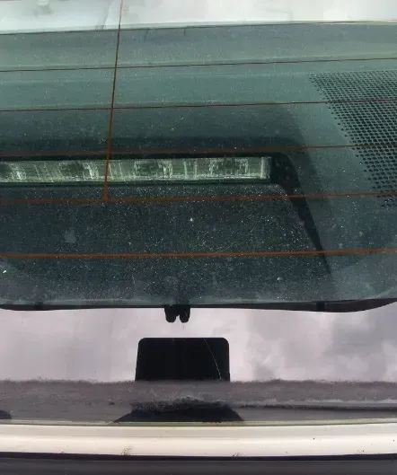 A close-up view through a car's rear windshield showing a horizontal high-mount brake light and rear window defroster lines.