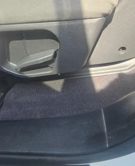 A grey plastic lever used to adjust a car seat, located on the lower side panel next to dark grey carpeted flooring.