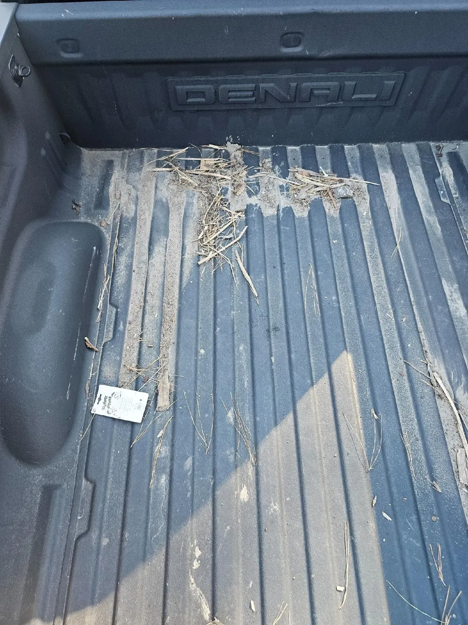 The interior bed of a GMC Denali pickup truck, covered in a light layer of dirt, debris, and a small scrap of paper.
