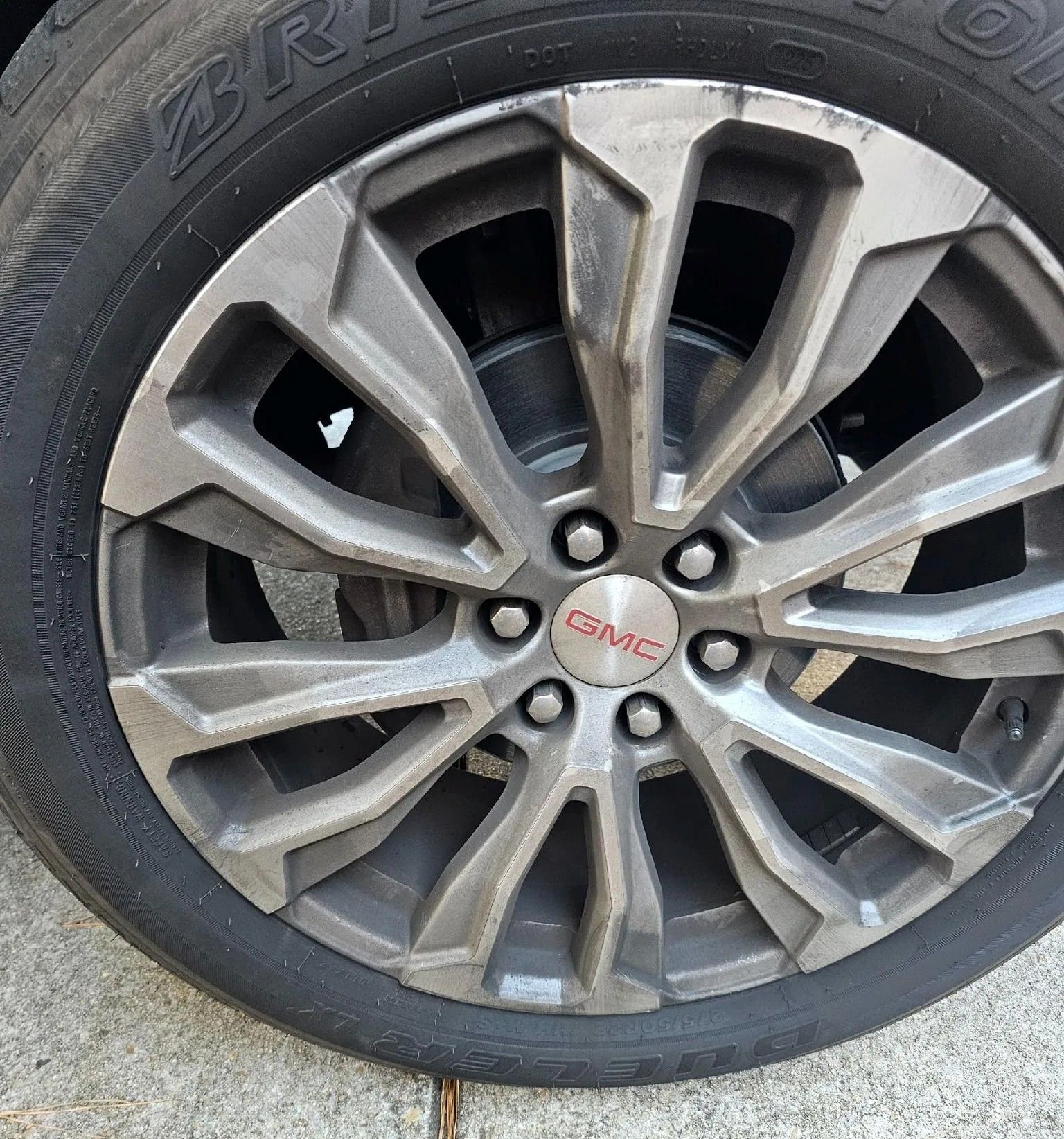 Close-up of a dark grey GMC multi-spoke alloy wheel with a Bridgestone tire, positioned on a concrete surface.