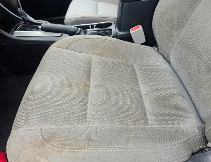 A light gray car seat with noticeable brown stains on the seat cushion fabric and near the side bolster.