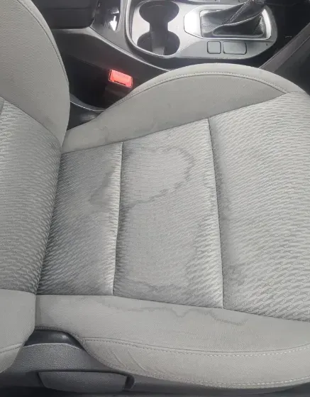A close-up view of a car's gray fabric seat with a large, wet, dark-colored stain across the center cushion.