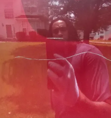 A person taking a selfie in a red-tinted, cracked mirror with a suburban home and trees visible in the background.