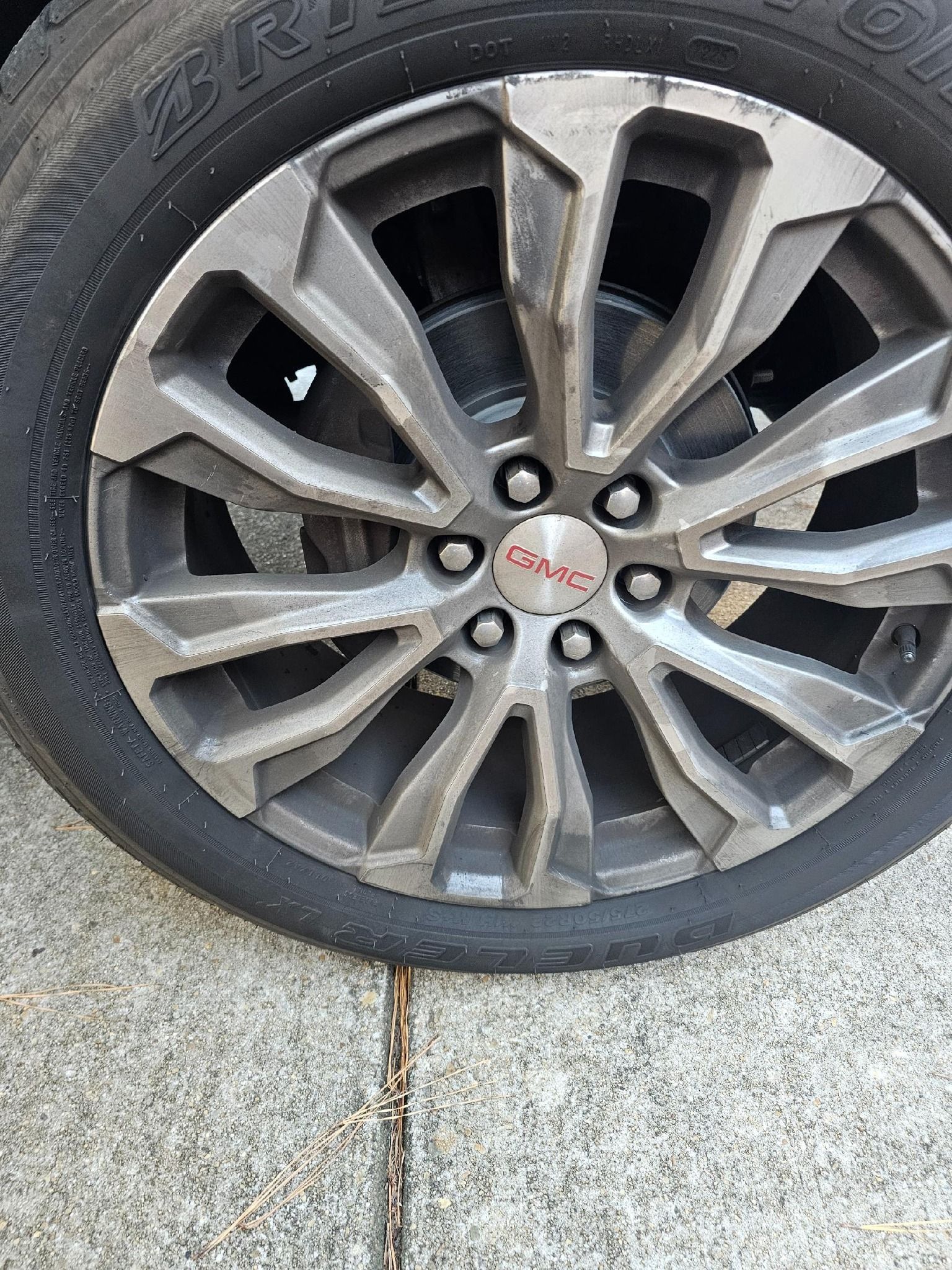 GMC wheel with tire on a concrete surface. Gray rim with red GMC logo in the center.