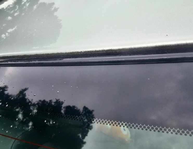 Close-up of a car's rear window, showing the top edge, seal, and the reflection of a cloudy sky and trees.